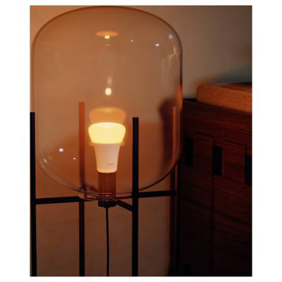A glowing smart bulb inside a transparent lamp on a stand, casting a warm light beside a wooden cabinet.