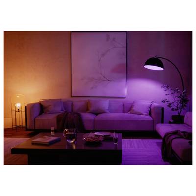 A cozy living room with a large, light purple couch, soft pillows, a curved floor lamp, and a warm ambient glow. Minimalist wall art.