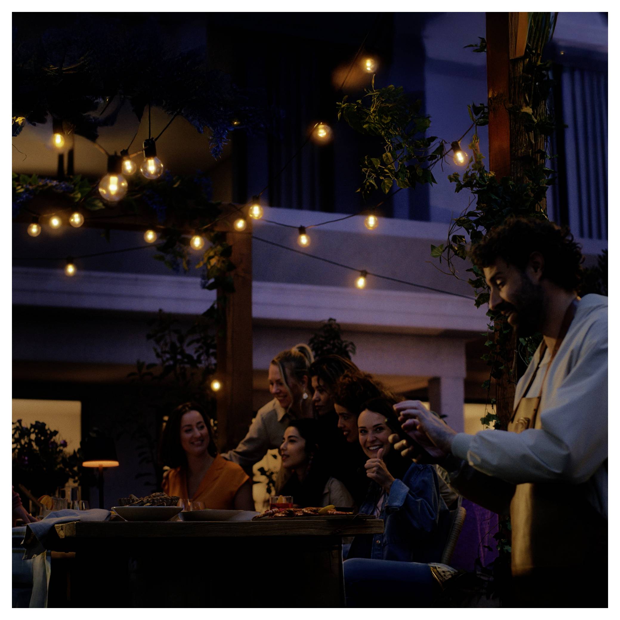A group of people enjoying an evening gathering under string lights, with a man pouring a drink and others smiling at a table.