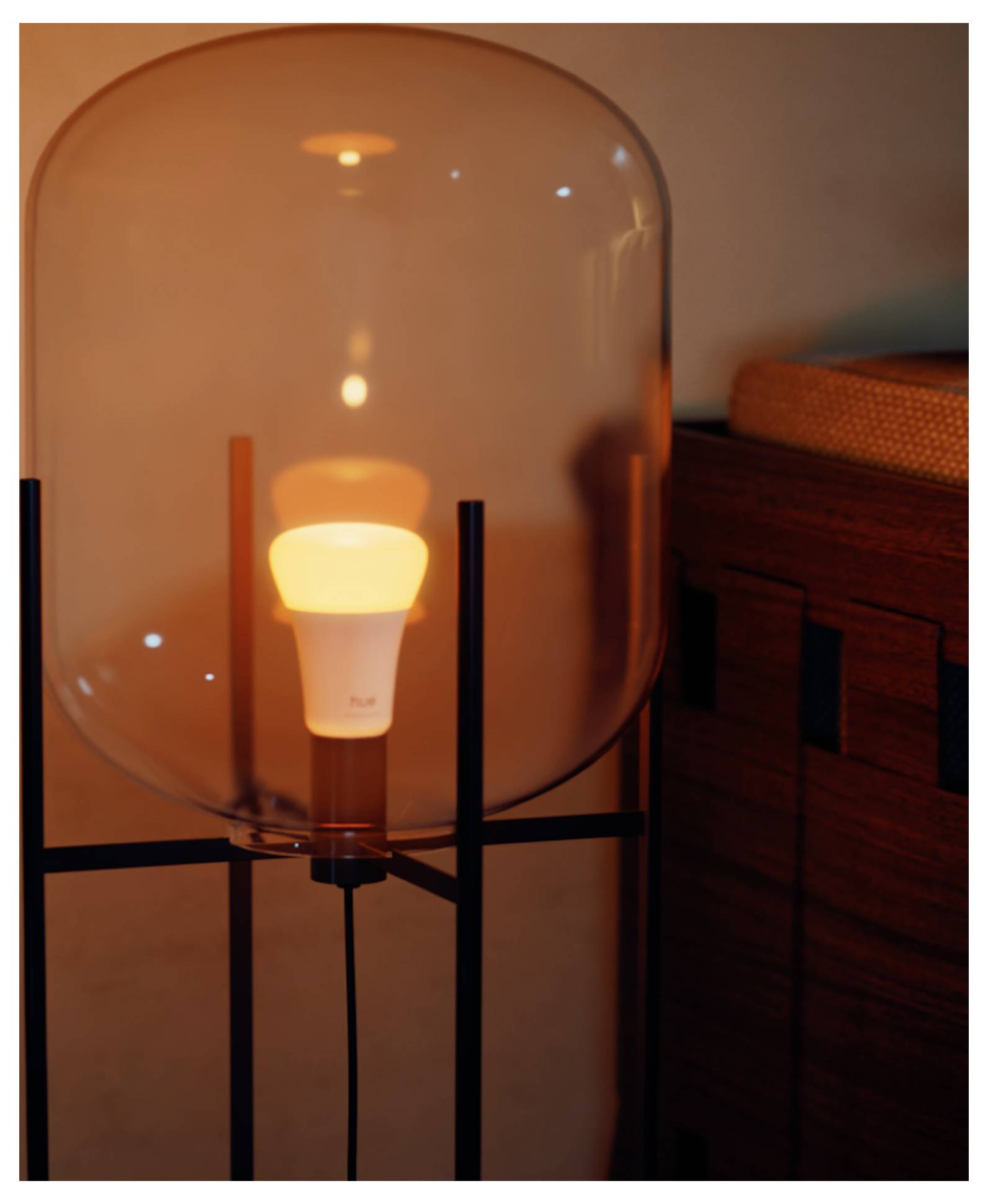 A glowing light bulb inside a modern, transparent glass lamp, placed beside a wooden cabinet in a softly lit room.