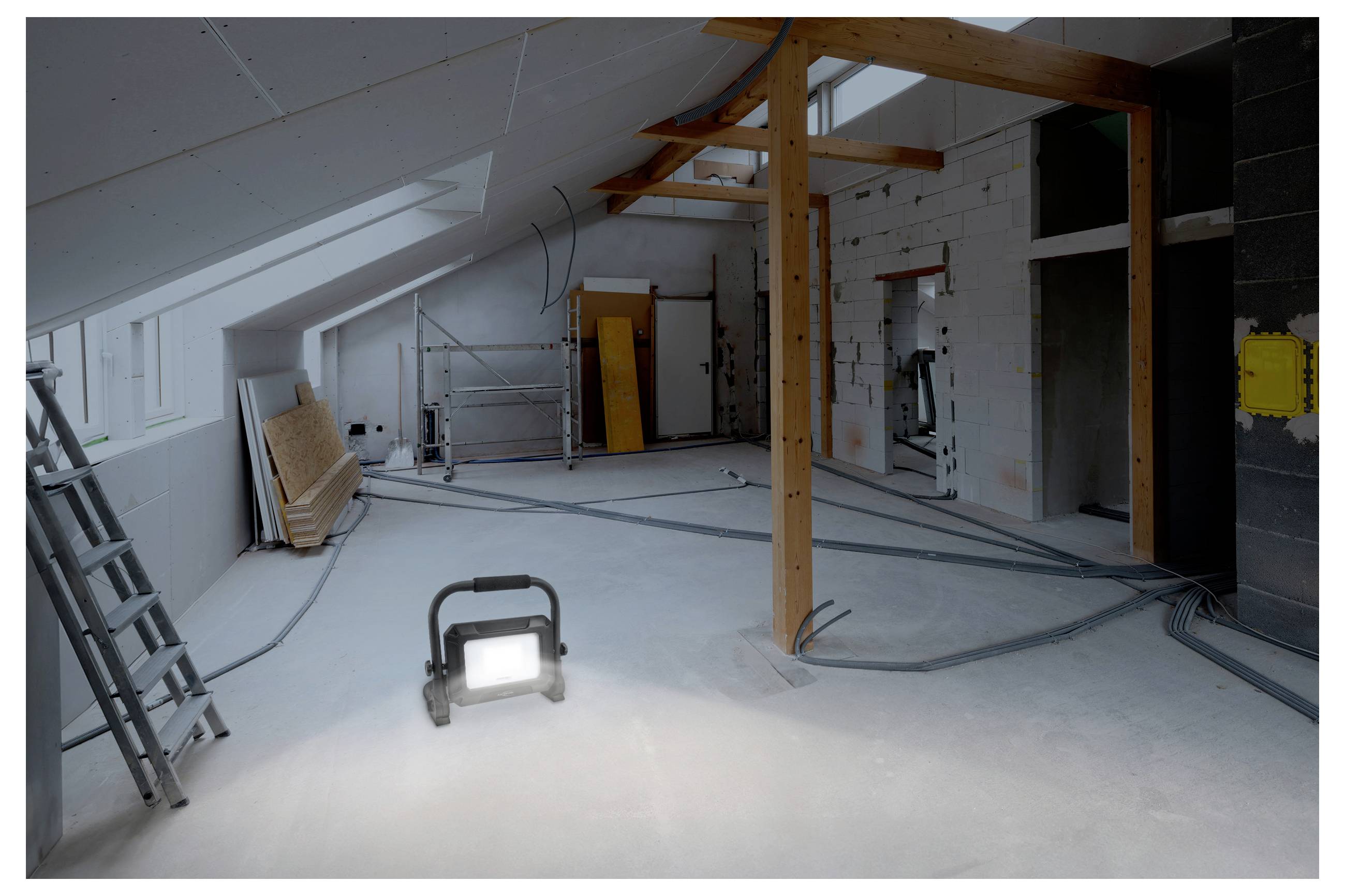 An unfinished attic room with exposed beams and wires, partial drywall, construction tools, and a work light on the floor.
