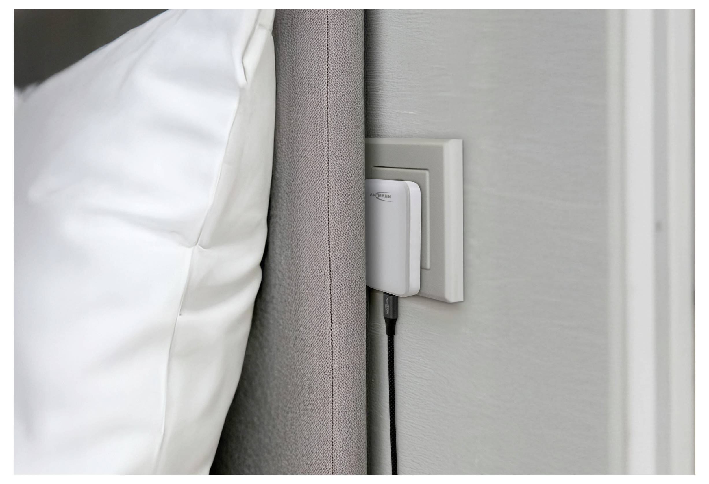 A white pillow leans against a gray padded headboard next to a wall socket with a plugged-in charger, indicating a bedroom setting.