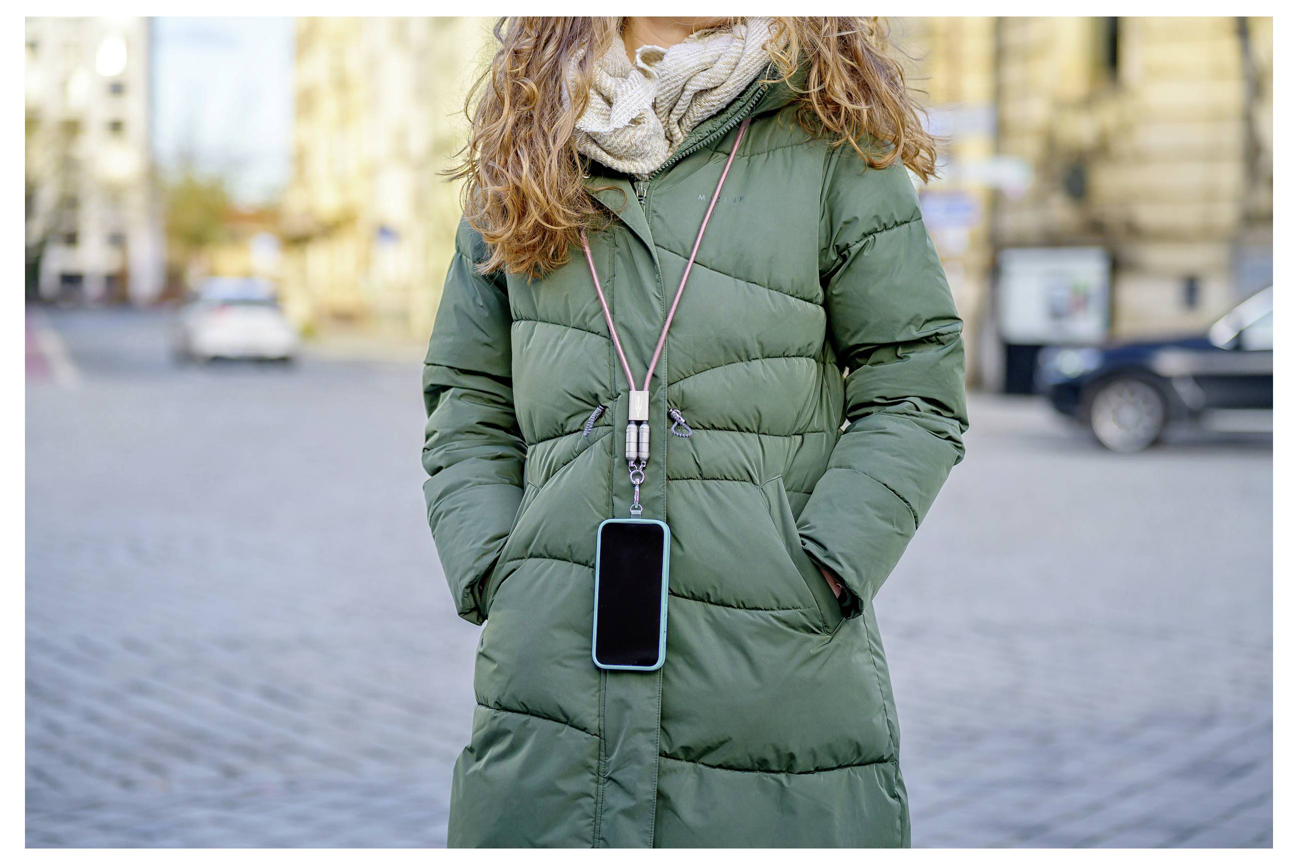A person in a green winter coat with a smartphone hanging from a lanyard around their neck on a city street.