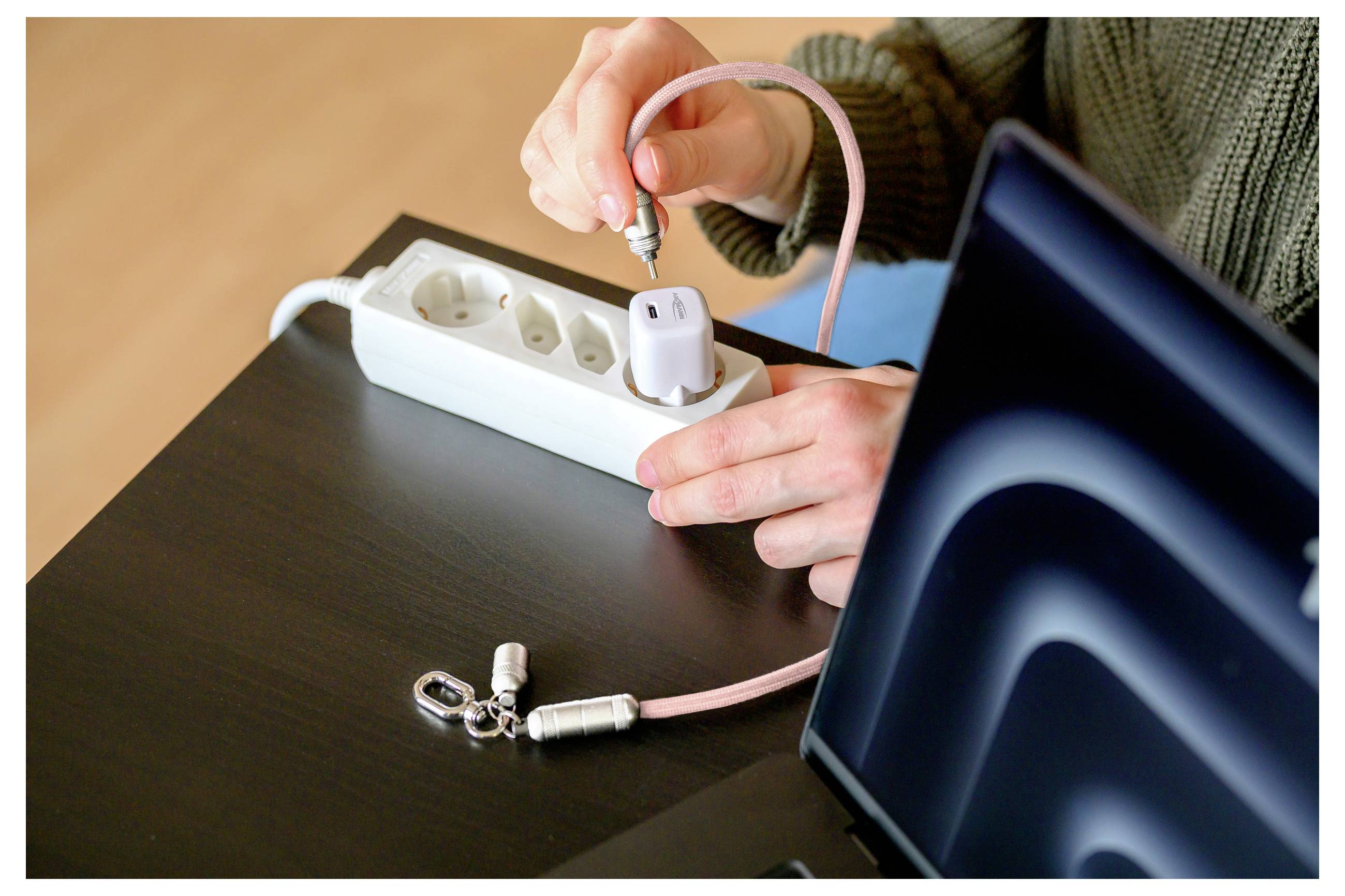 A person connects a pink charging cable to a white power strip on a dark table beside a laptop, preparing to charge a device.