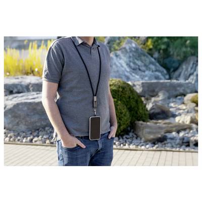 A man in a gray polo shirt and jeans stands outdoors with his hands in pockets, wearing a large smartphone on a lanyard around his neck.