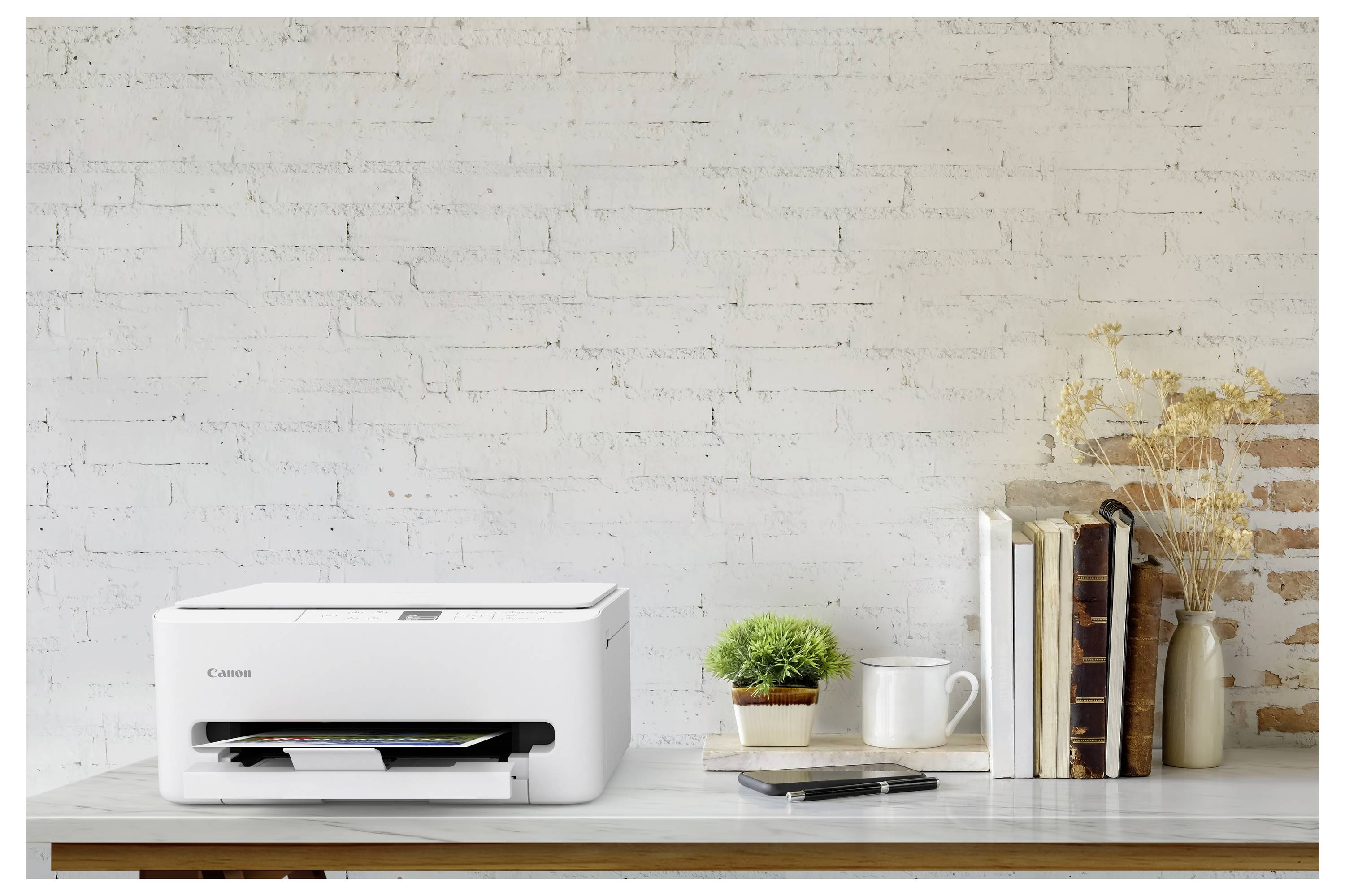 A compact white printer on a wooden desk next to a small plant, books, and a smartphone, with a white brick wall in the background.