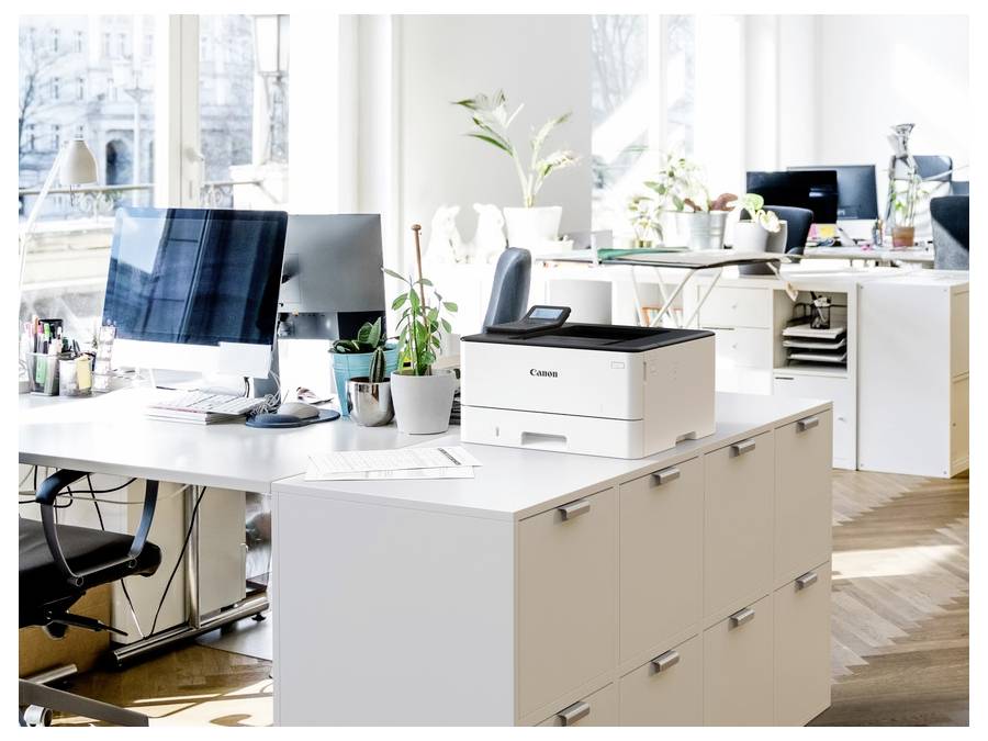 An open, modern office space with desks, computers, and a Canon printer on a cabinet. Large windows provide natural light and city views.