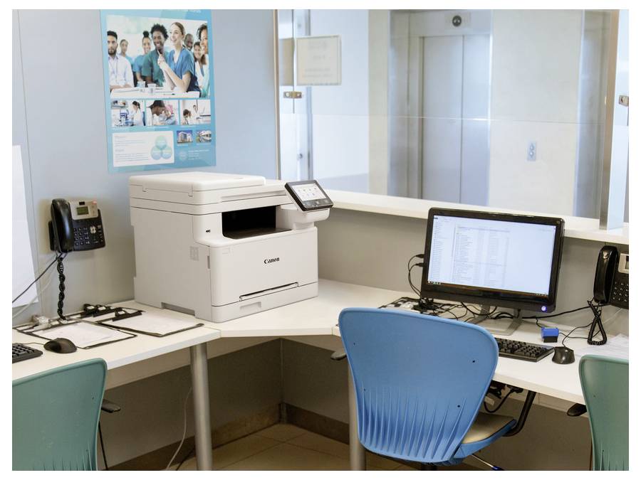 The image shows an office setup with a white printer, a computer with a blue chair, and telephones on a desk. Posters are on the wall.