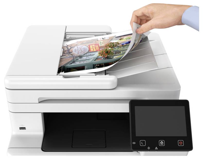 A hand places a photo into a white multifunction printer/copier with a control panel. The printer lid is open, showing the photo being scanned.