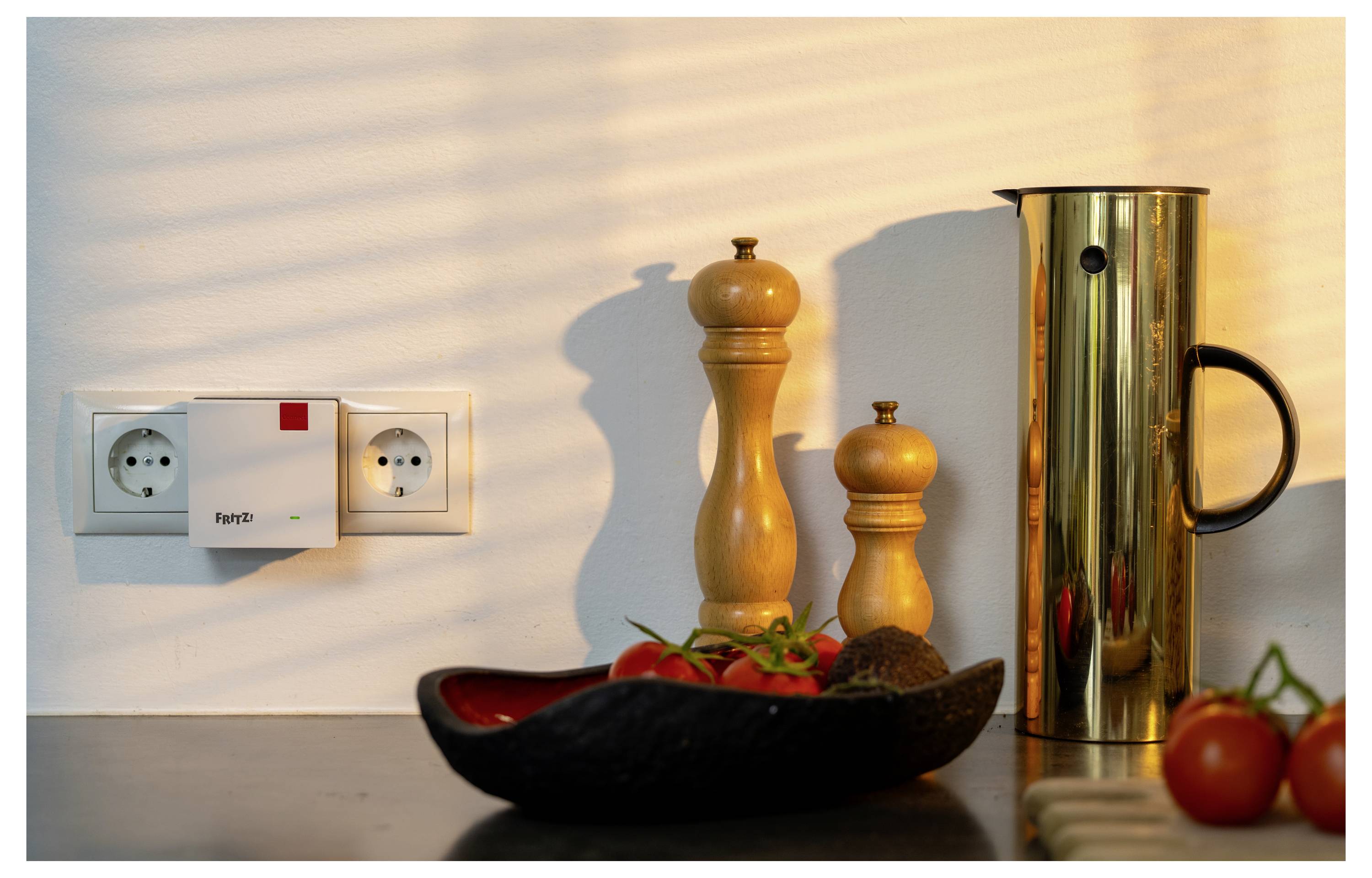 Kitchen counter with a plugged-in gas detector, pepper mills, a gold pitcher, and a bowl of tomatoes and avocados, lit by sunlight.