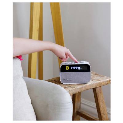 A hand presses a button on a small speaker displaying 'Pairing...' on the screen, indicating a Bluetooth connection attempt.