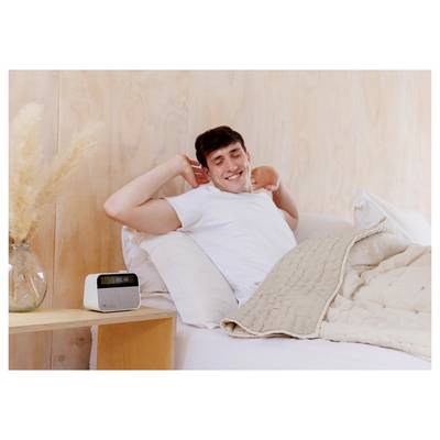 A person in bed stretches with a smile, next to a nightstand with an alarm clock reading '7:00 AM'. The room is softly lit.