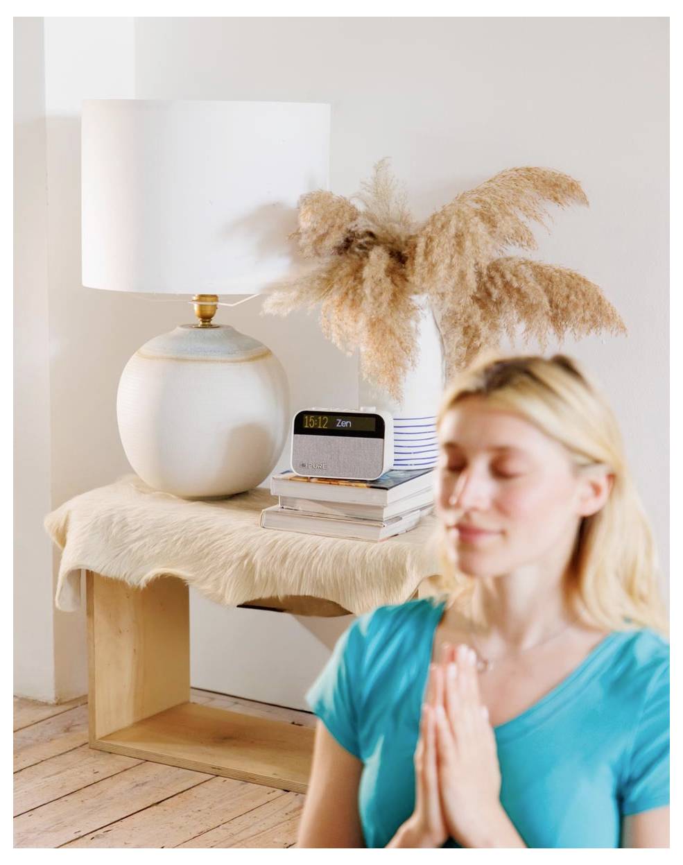A person in a blue shirt meditates with hands together in prayer position. Behind, a clock, books, lamp, and pampas grass on a table.