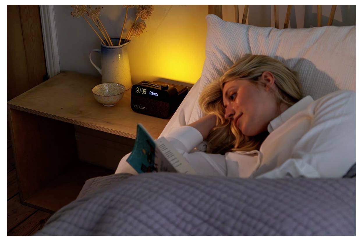 A woman lies in bed under a purple quilt, reading a book. A warm light glows beside her on a wooden nightstand, illuminating the space.