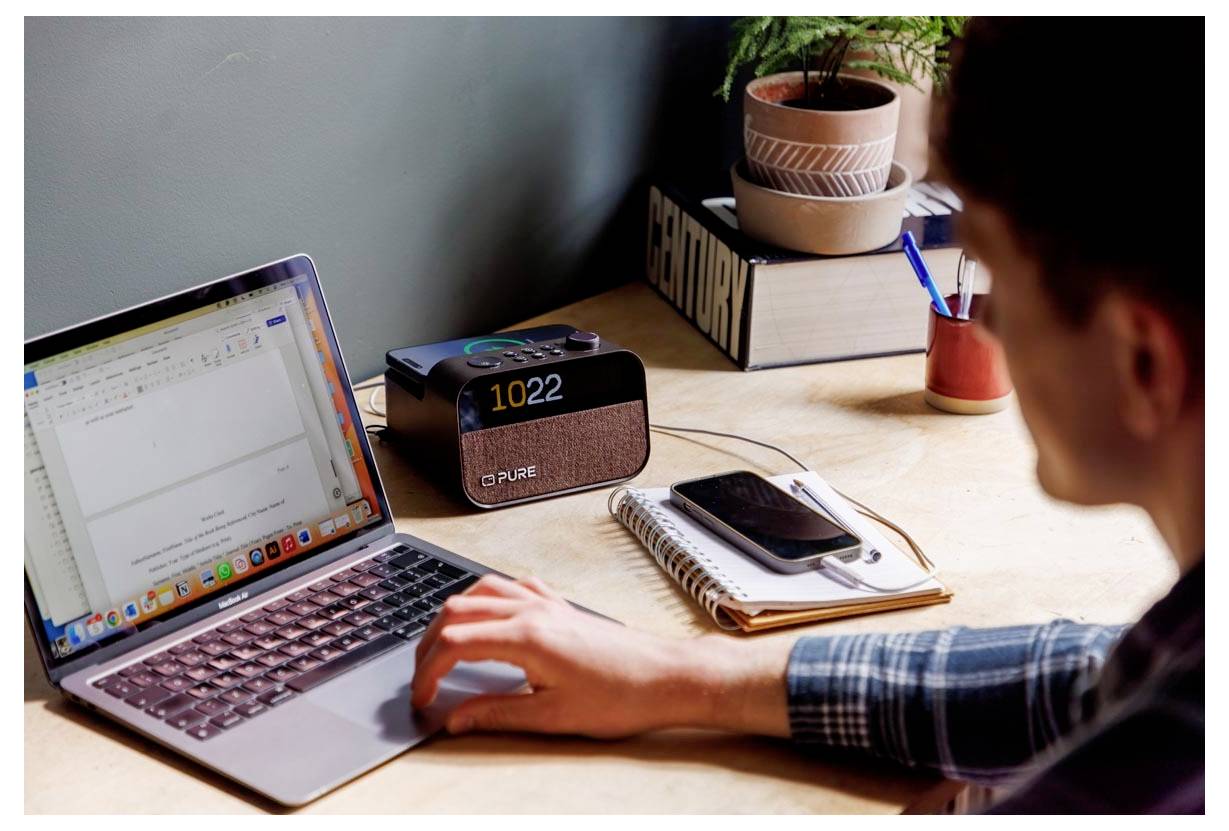 A person types on a laptop at a desk, with a digital clock showing '10:22', a plant, and a stack of books in the background.
