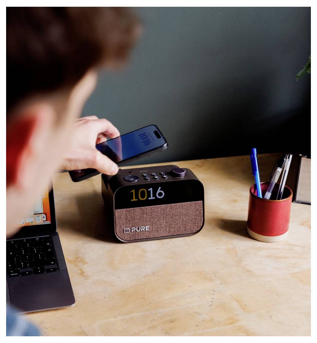 A person holds a smartphone near a digital clock radio displaying '10:16'. Nearby are a laptop, a pen holder, and a small plant.