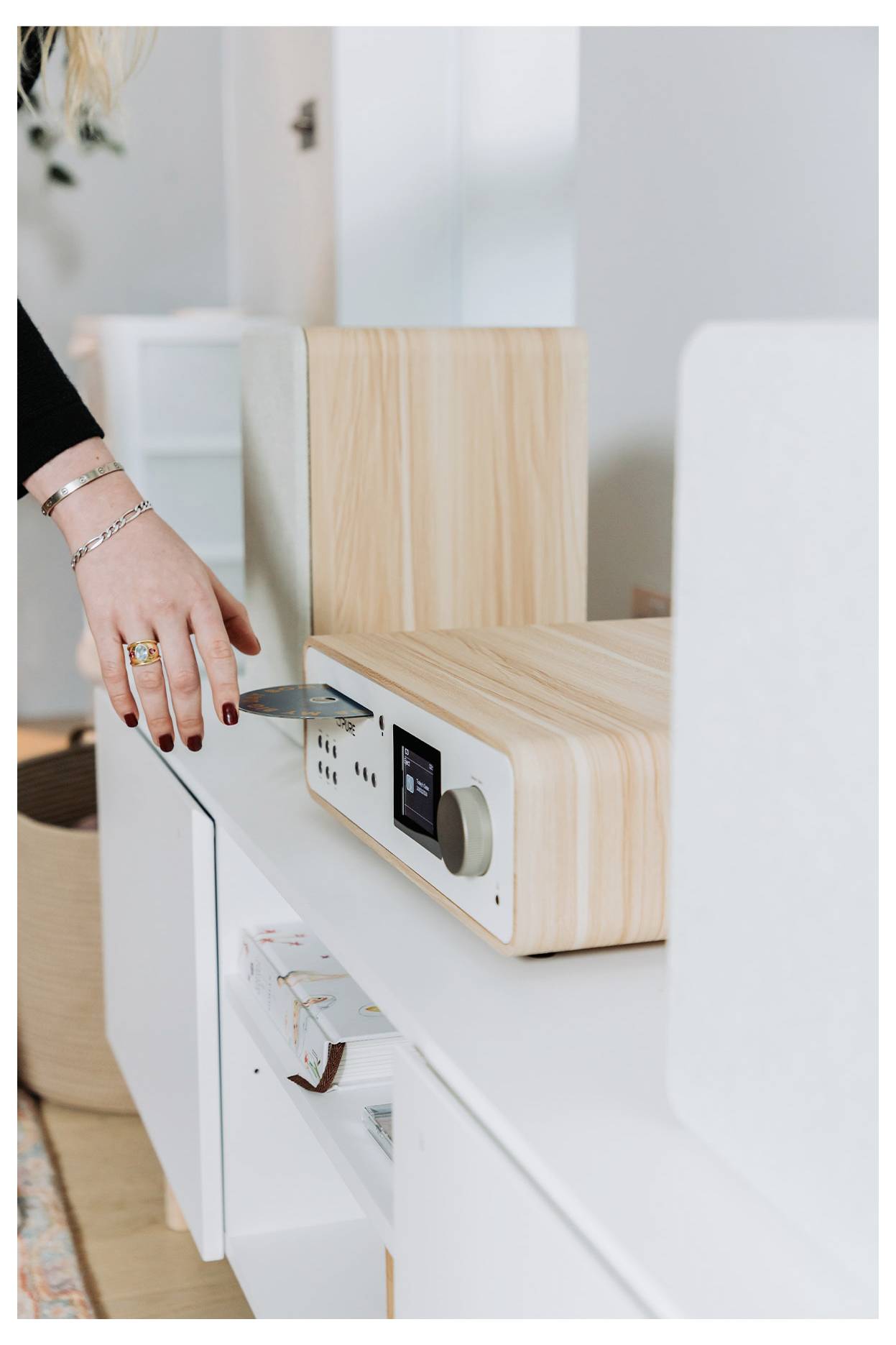 A person is inserting a CD into a modern wooden stereo system on a white shelf, with books and decor visible in the background.