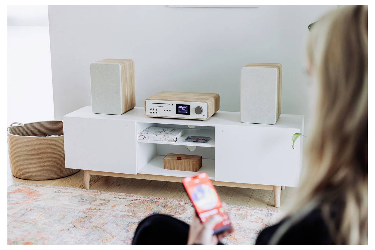 A person holds a smartphone displaying a music app. In the background, a modern stereo system with speakers is set up on a shelf.