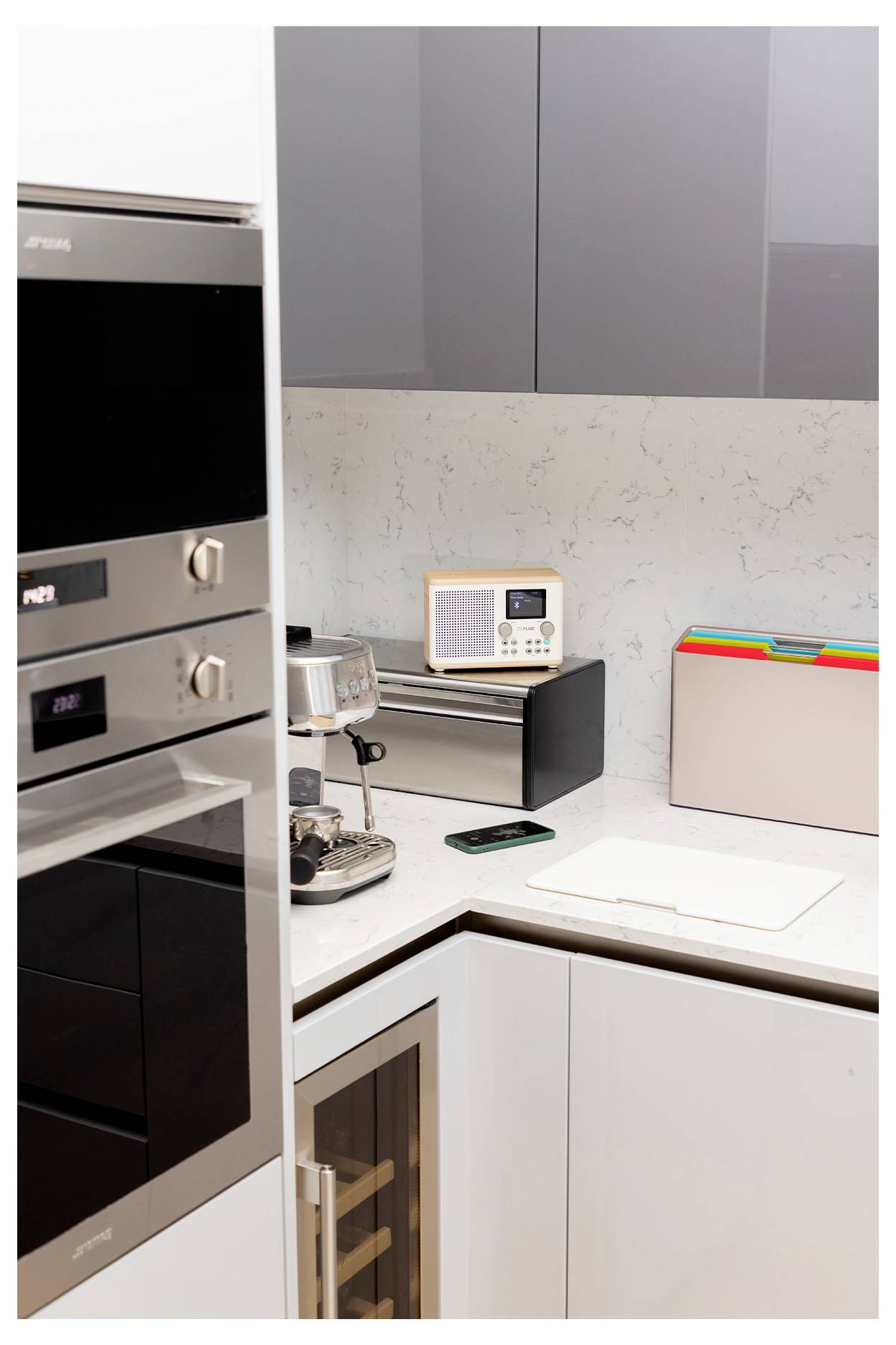 Modern kitchen corner with stainless steel oven, coffee machine, radio, and bread box on marble countertop. Grey and white cabinets.