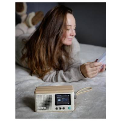 A woman lying on a bed reads a paper. In the foreground, a retro-style radio displays a small screen and control buttons.