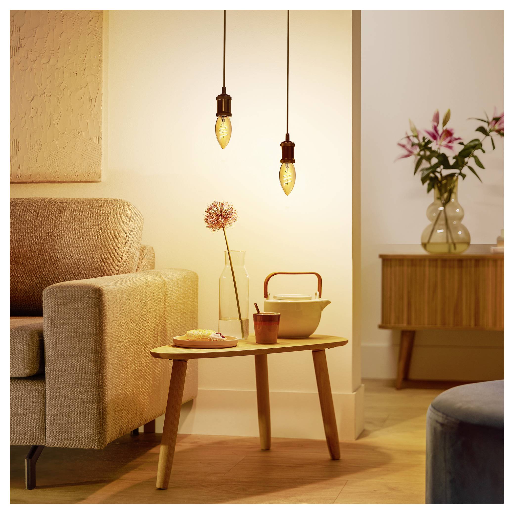 A cozy living room corner with a beige armchair, wooden table, vase with pink flowers, and two hanging light bulbs, creating a warm ambiance.