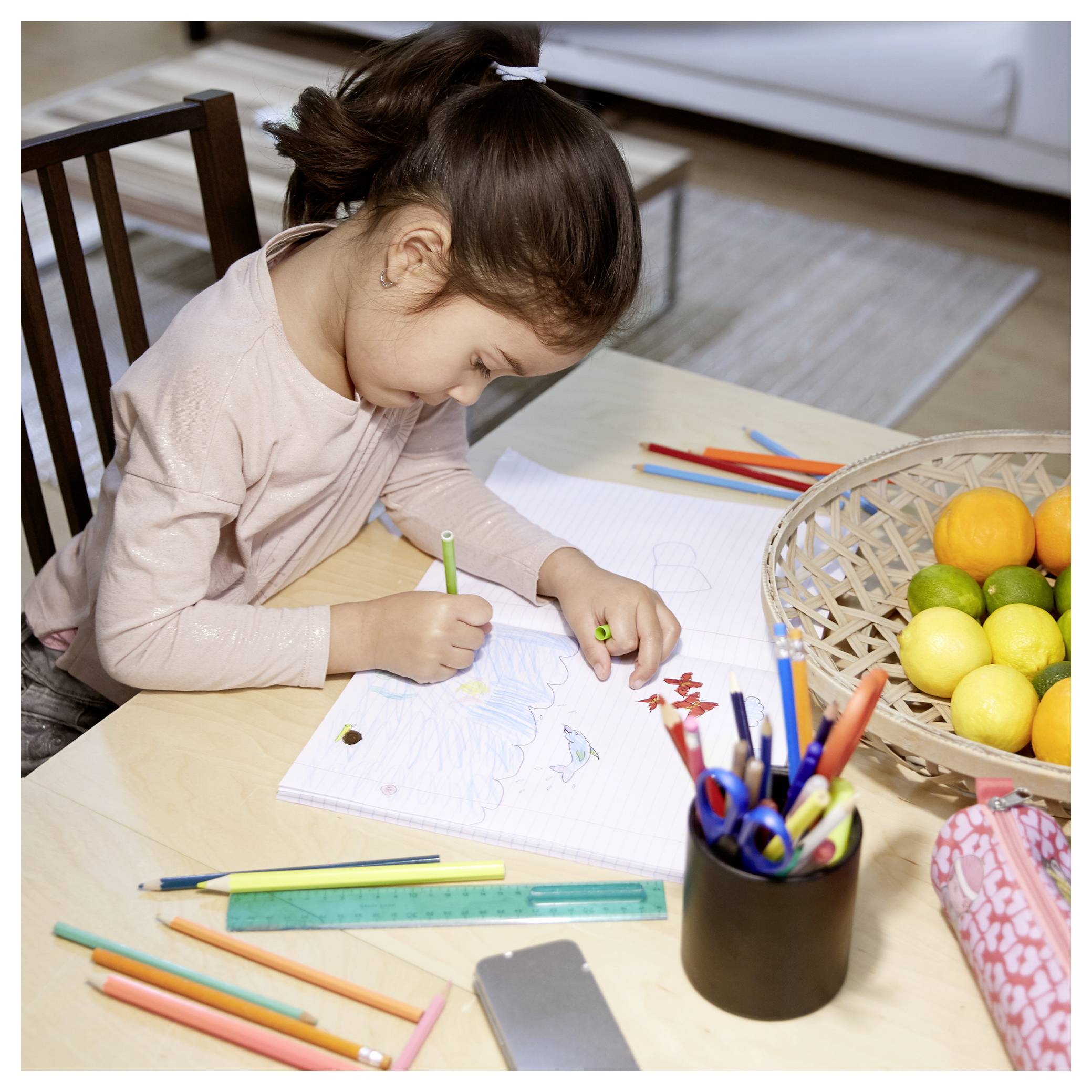 A young child drawing with colored pencils at a table, surrounded by drawing materials and a bowl of fruit.