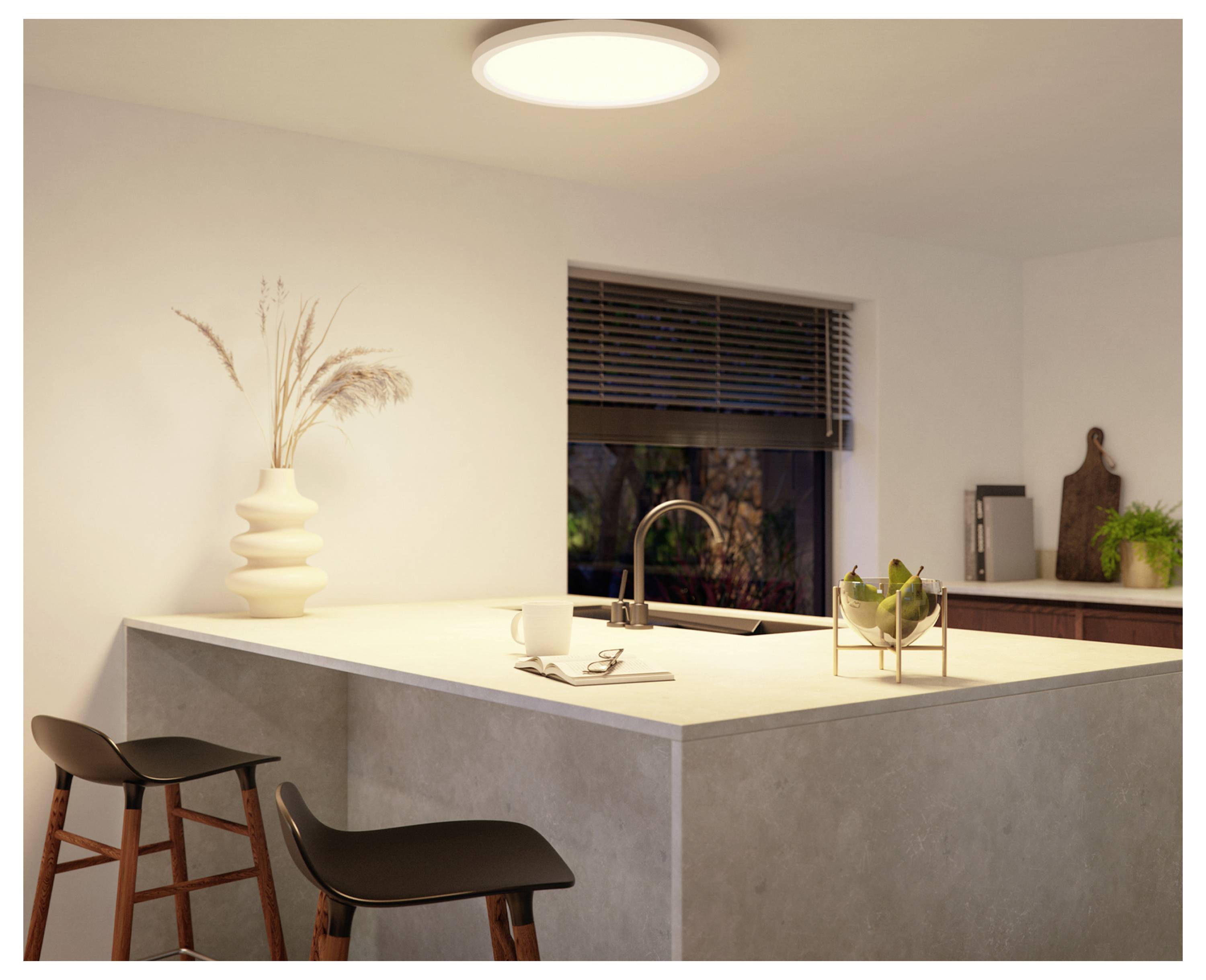 A modern kitchen with a corner countertop featuring two stools, a vase with dried plants, a faucet, and a bowl of green apples.