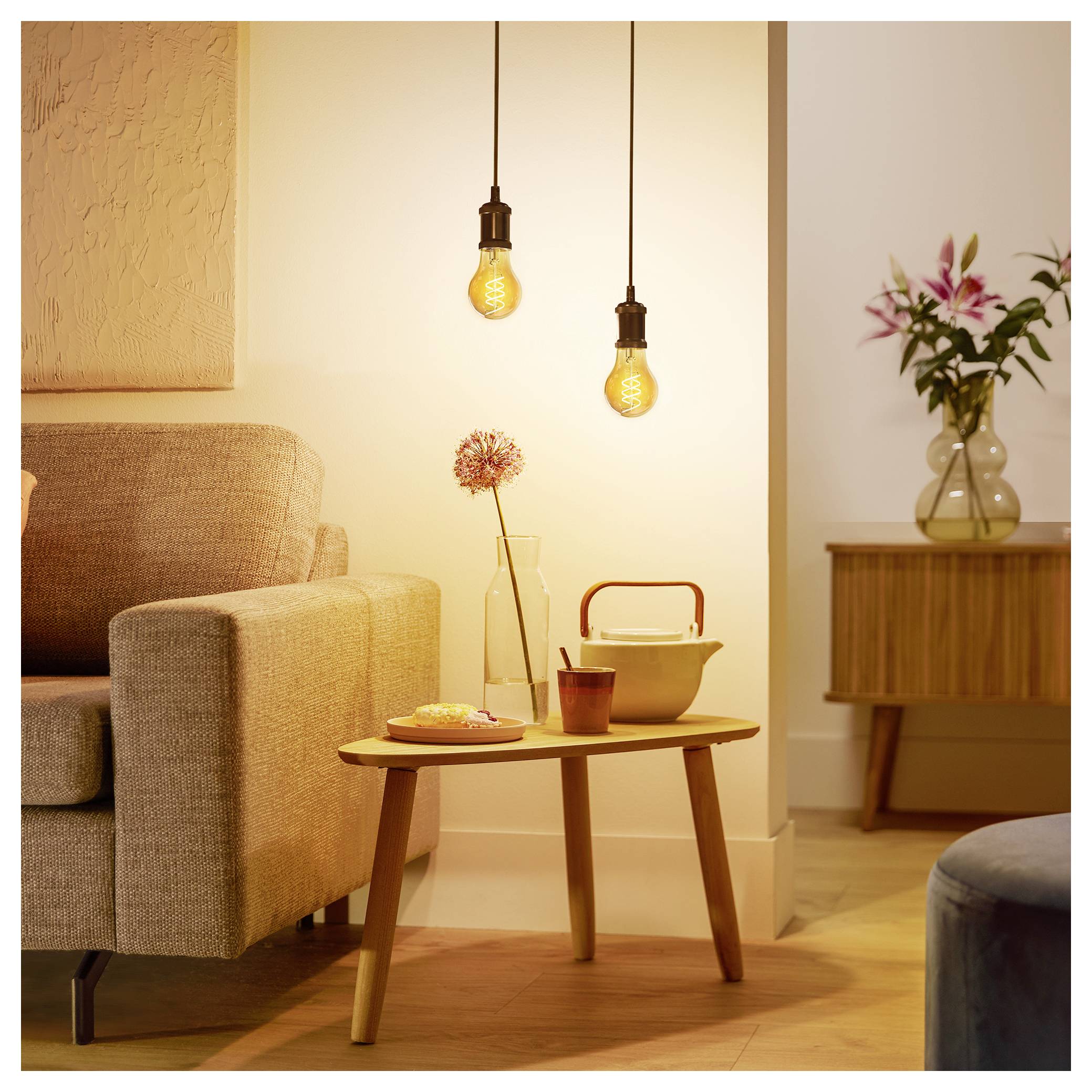 A cozy living room with warm lighting, featuring a beige sofa, wooden coffee table with teapot and cup, and two hanging light bulbs.