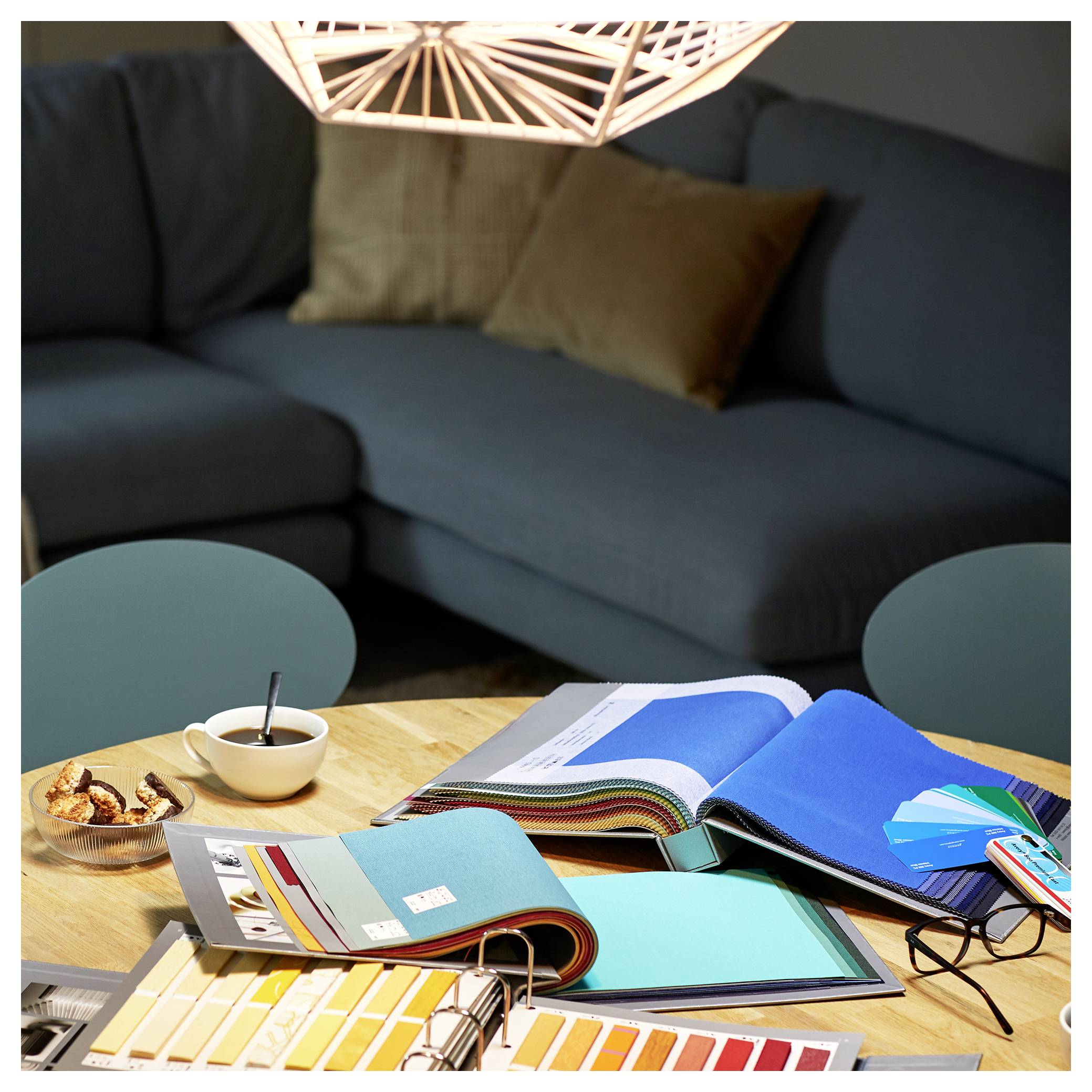 A table with open color sample books, fabric swatches, and design materials under a modern light, with a sofa in the background.