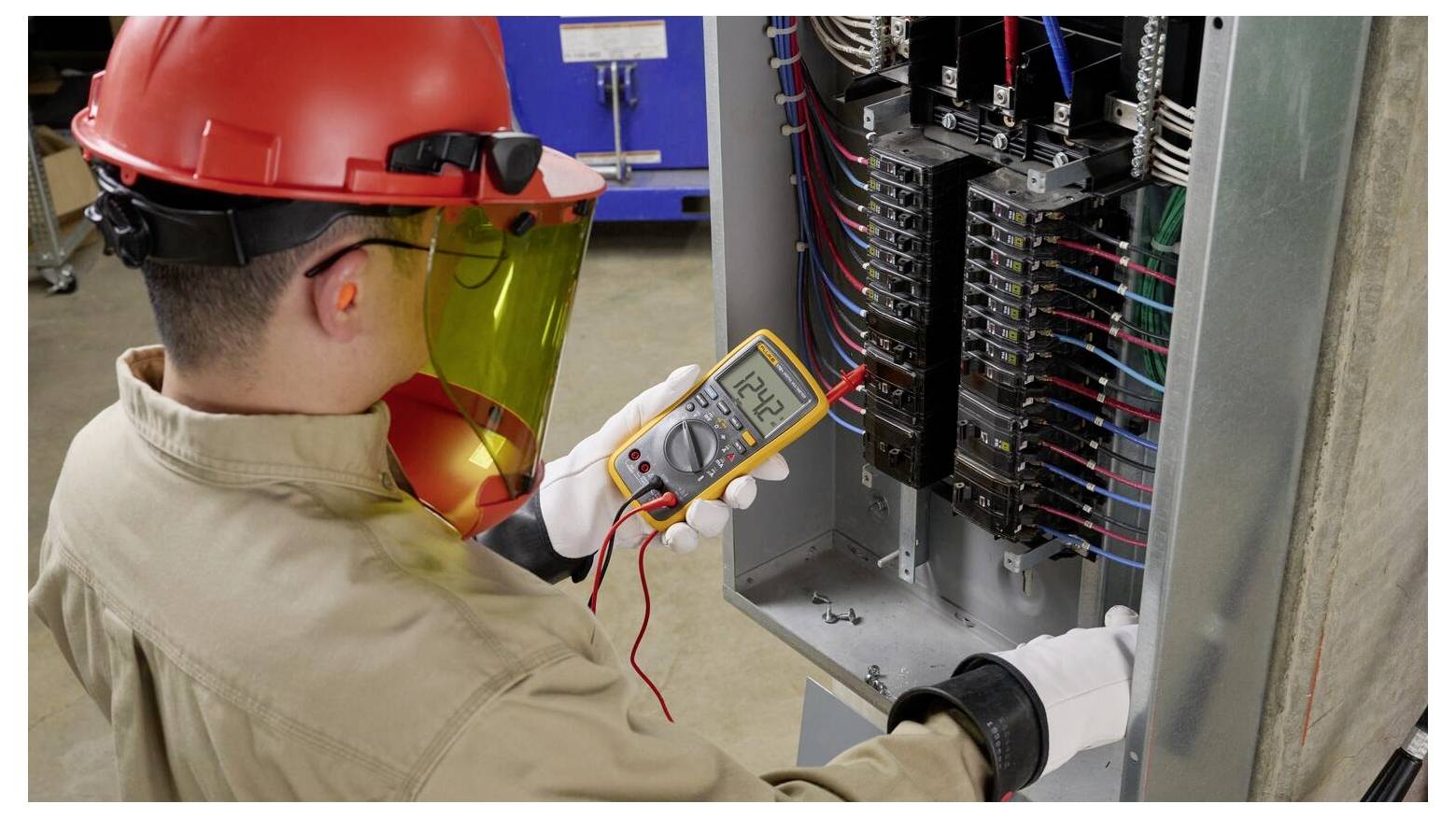 A worker wearing protective gear uses a multimeter to check electrical wiring inside an open panel, indicating a safety inspection.