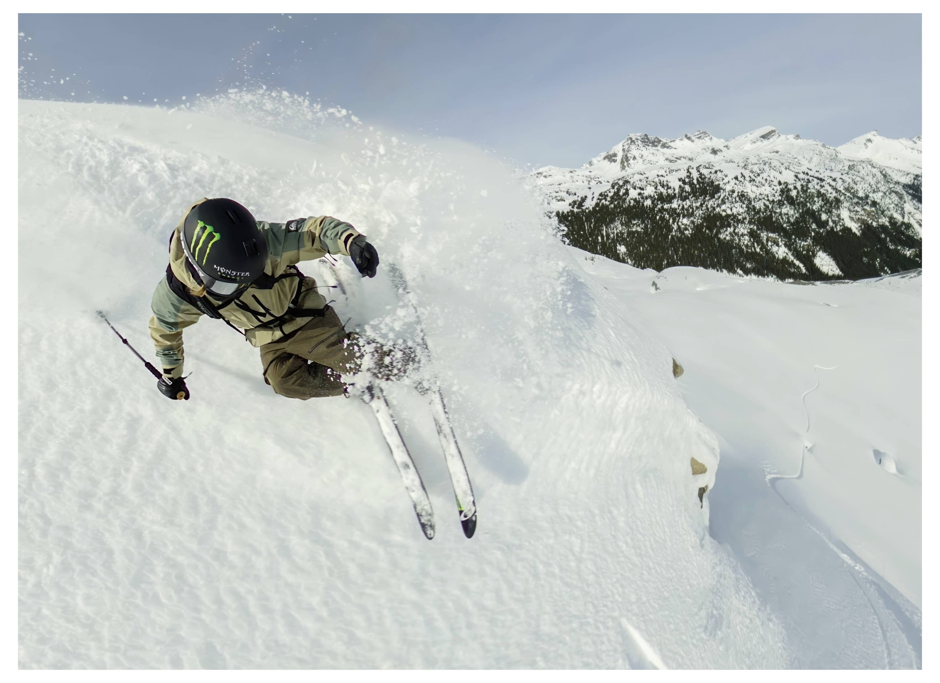 A skier performs a daring downhill maneuver on a snowy mountain slope, surrounded by vast snow-covered peaks and a clear sky.