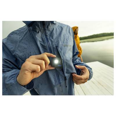 A person in a rain jacket holds a small, illuminated LED device close to the chest pocket. Another person in a yellow jacket stands near water.