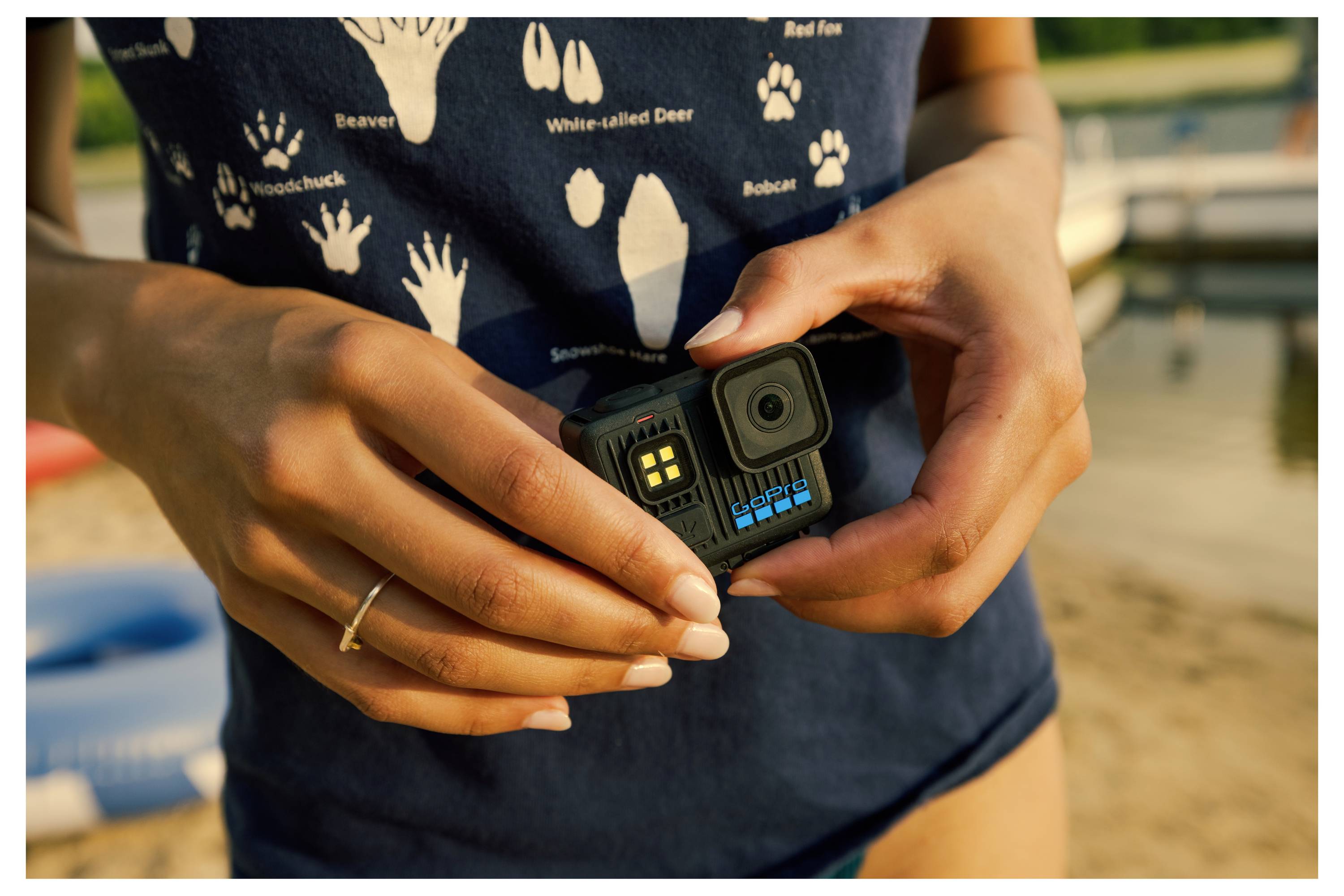 A person holds a compact action camera with a lake and boats in the background, wearing a T-shirt featuring animal tracks.