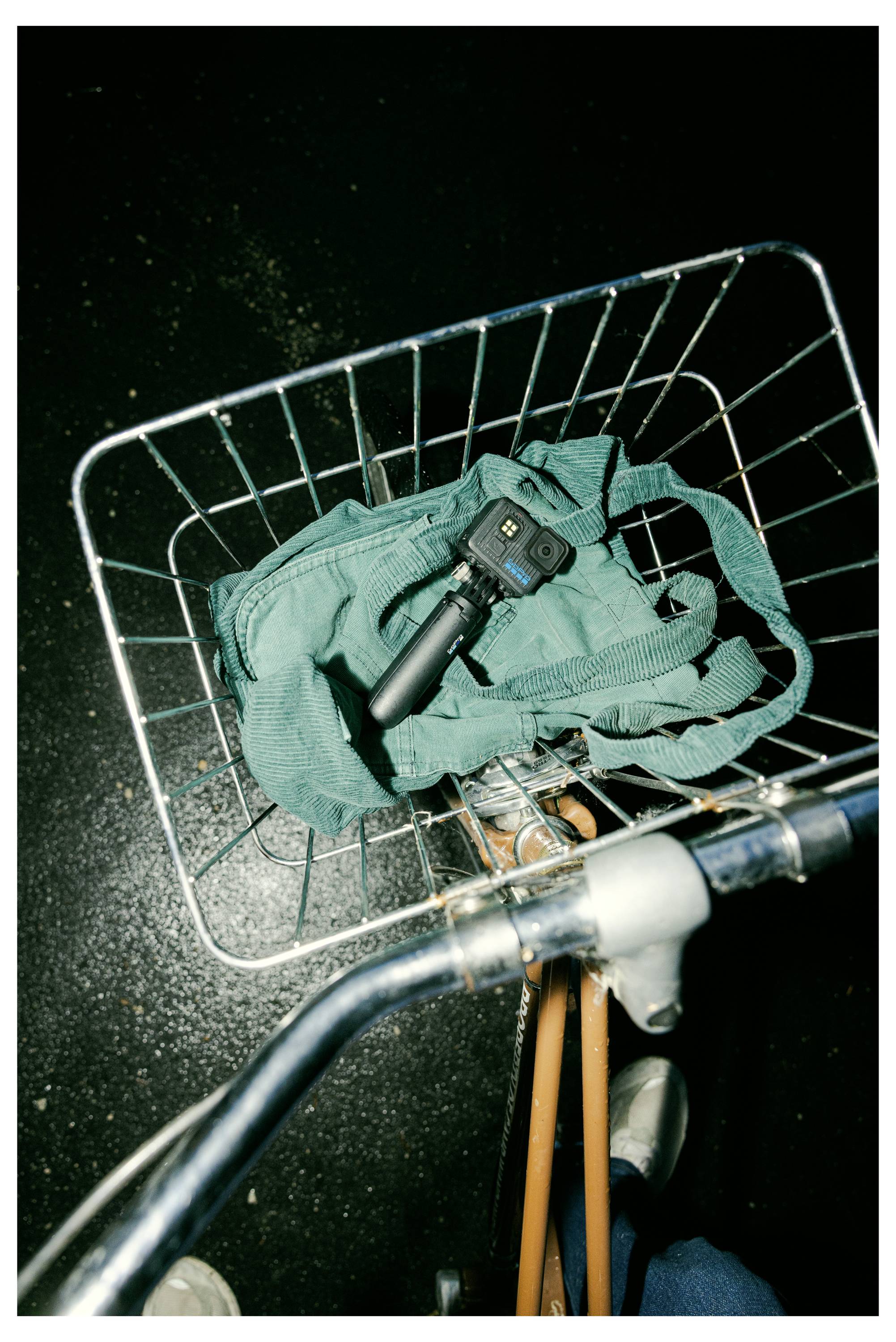 A bicycle basket holding a green bag and a handheld camera device with a gimbal, seen from above on a dimly lit surface.