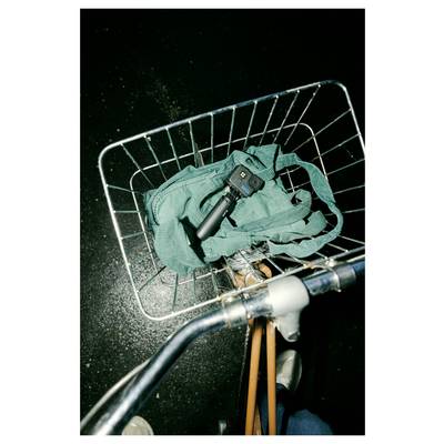 A bicycle basket holding a green bag and a handheld camera device with a gimbal, seen from above on a dimly lit surface.