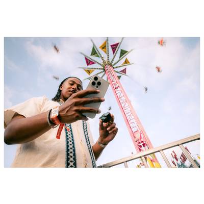 A person using a smartphone stands in front of a tall, colorful amusement ride with people swinging in motion against a blue sky.