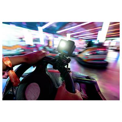 A person drives a bumper car in an indoor amusement setting, holding a steering wheel with a camera mounted on it. Bright lights blur in motion.