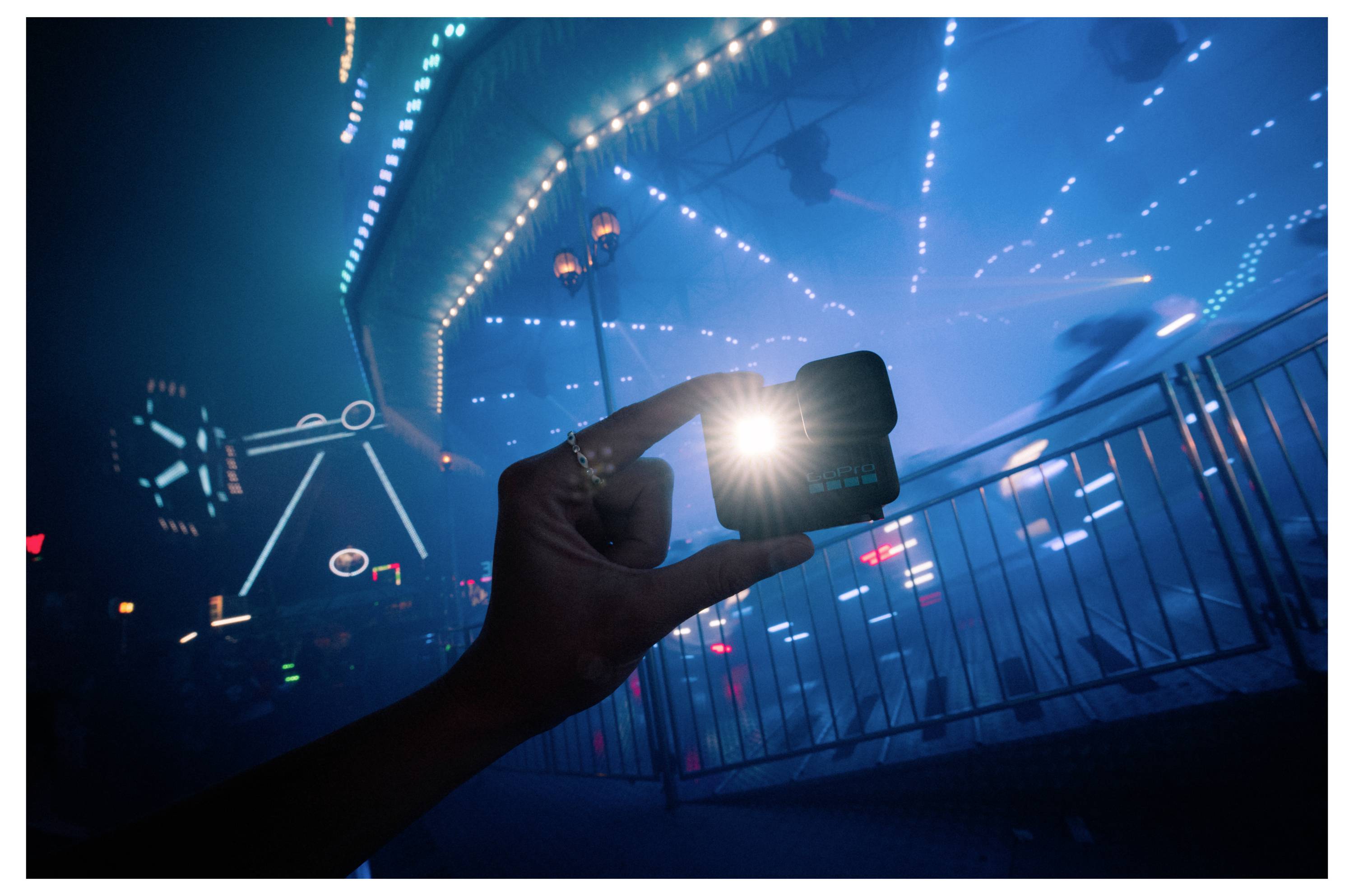 A hand holding a small, lit device against a backdrop of a blurred, illuminated carnival ride at night, creating a vibrant, dynamic scene.