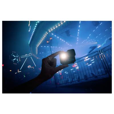 A hand holding a small, lit device against a backdrop of a blurred, illuminated carnival ride at night, creating a vibrant, dynamic scene.