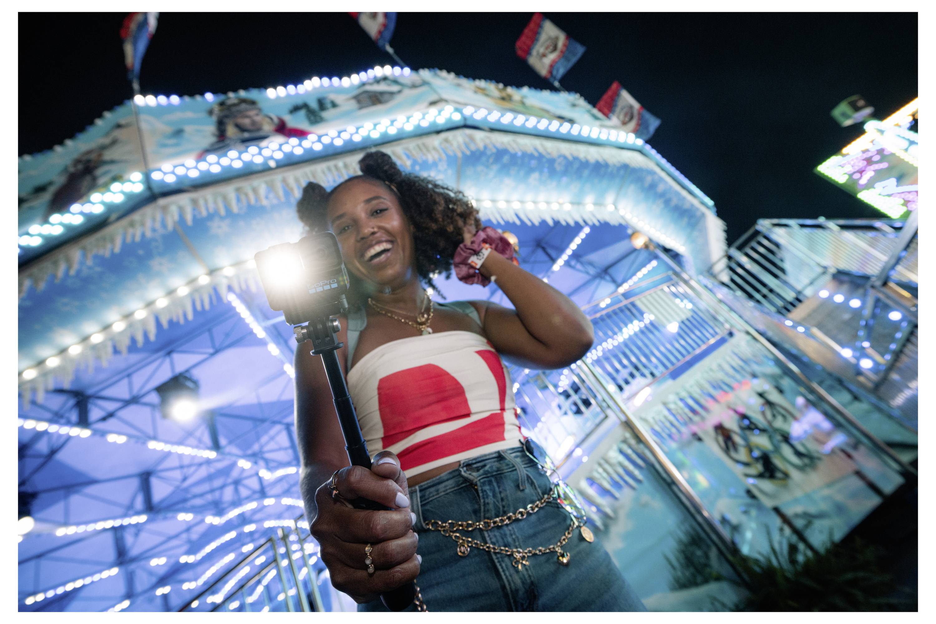 A person smiles and holds a camera on a stick at a brightly lit amusement park, with colorful rides and a lively atmosphere in the background.