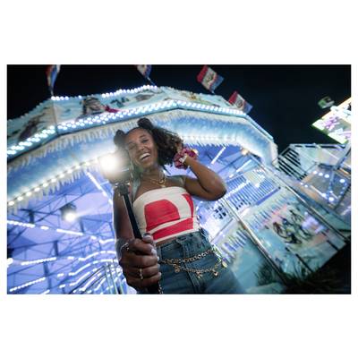 A person smiles and holds a camera on a stick at a brightly lit amusement park, with colorful rides and a lively atmosphere in the background.