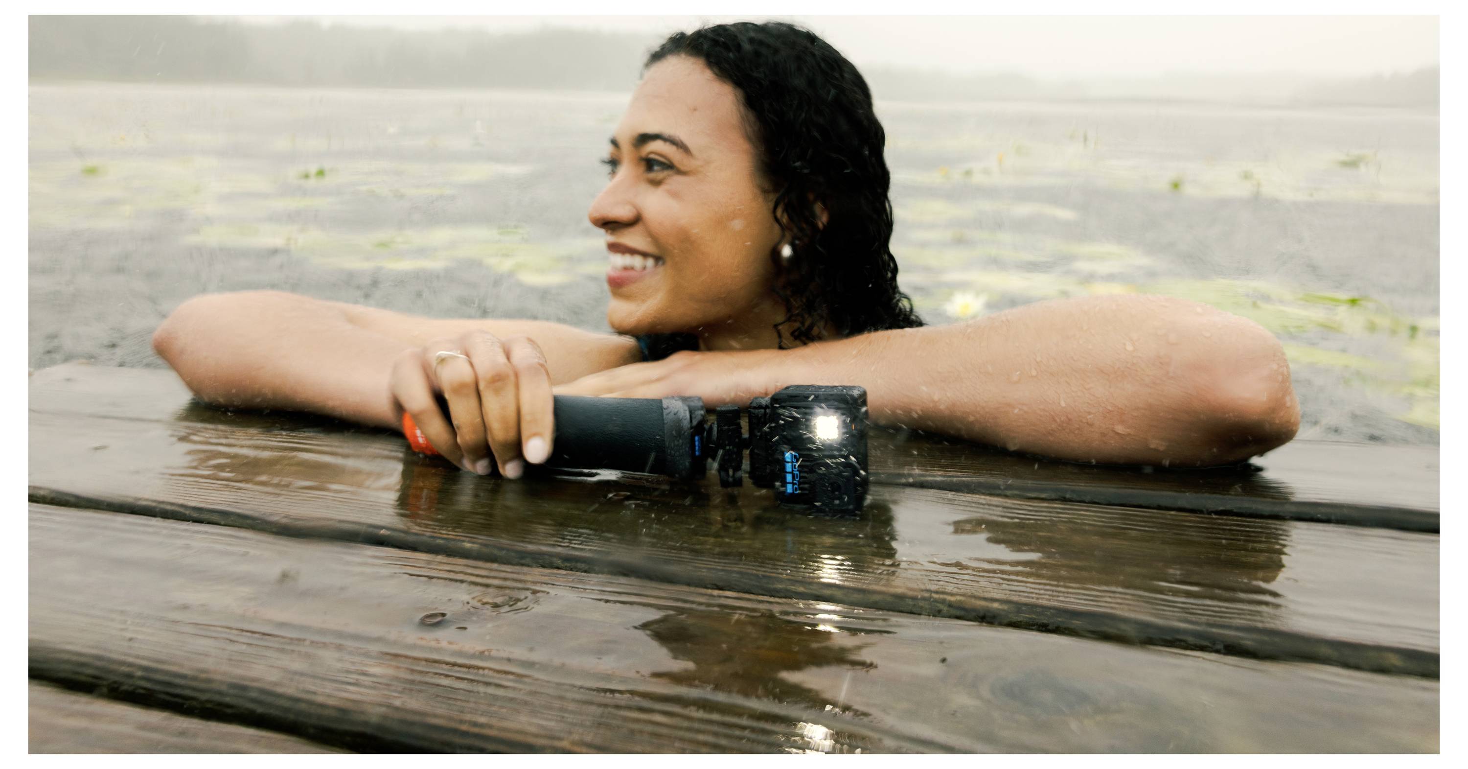 A person with curly hair smiling, partially submerged in water while resting their arms on a wooden dock, holding a small flashlight.