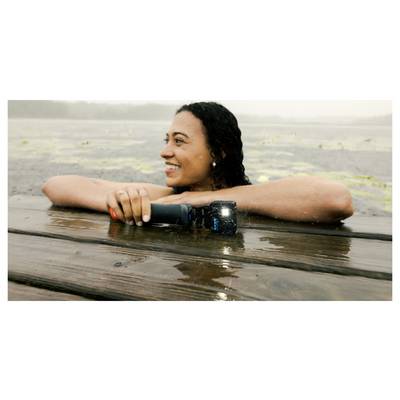 A person with curly hair smiling, partially submerged in water while resting their arms on a wooden dock, holding a small flashlight.