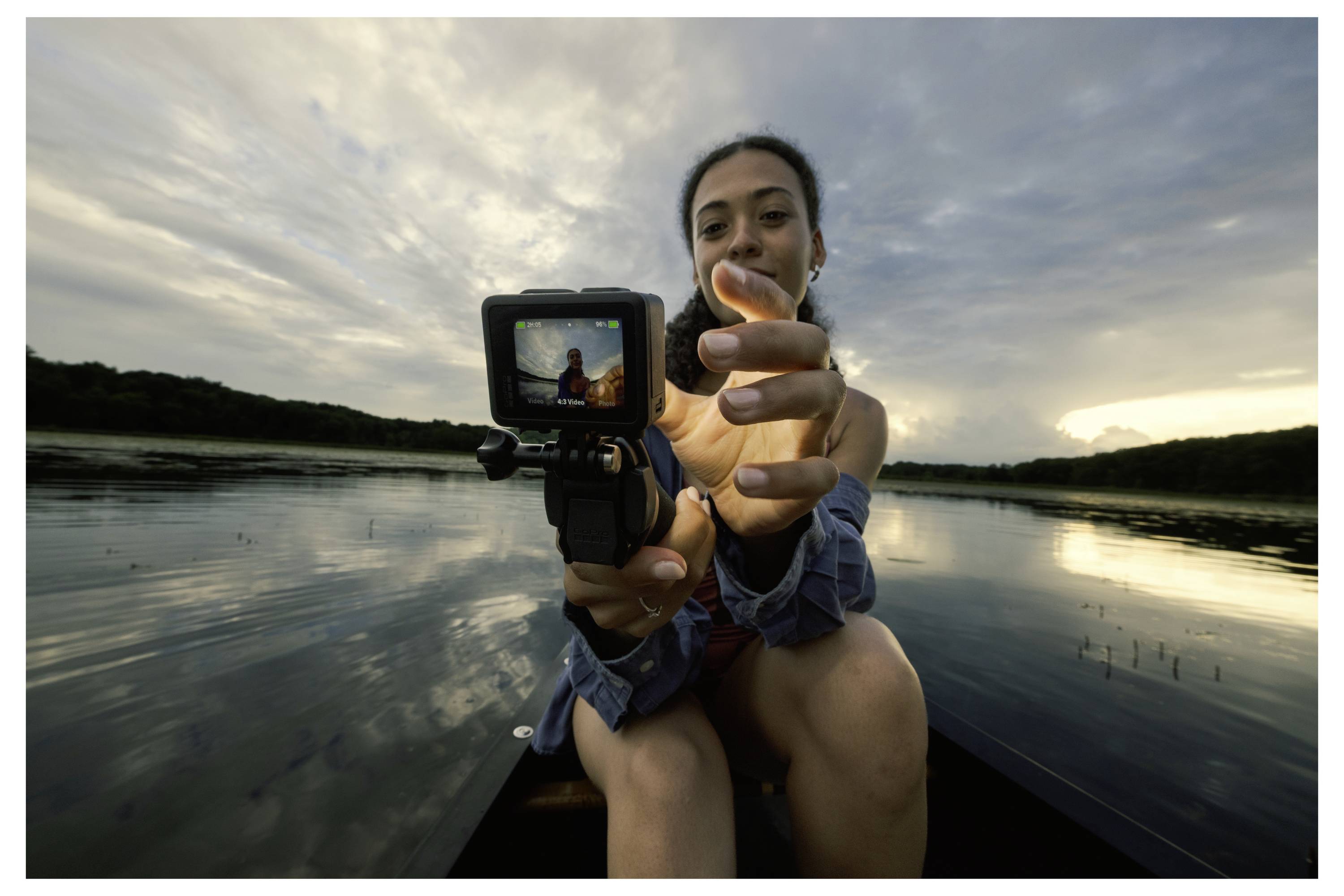 A person in a boat holds a camera, showing a view of the lake and cloudy sky behind. The scene is captured on the camera's screen.