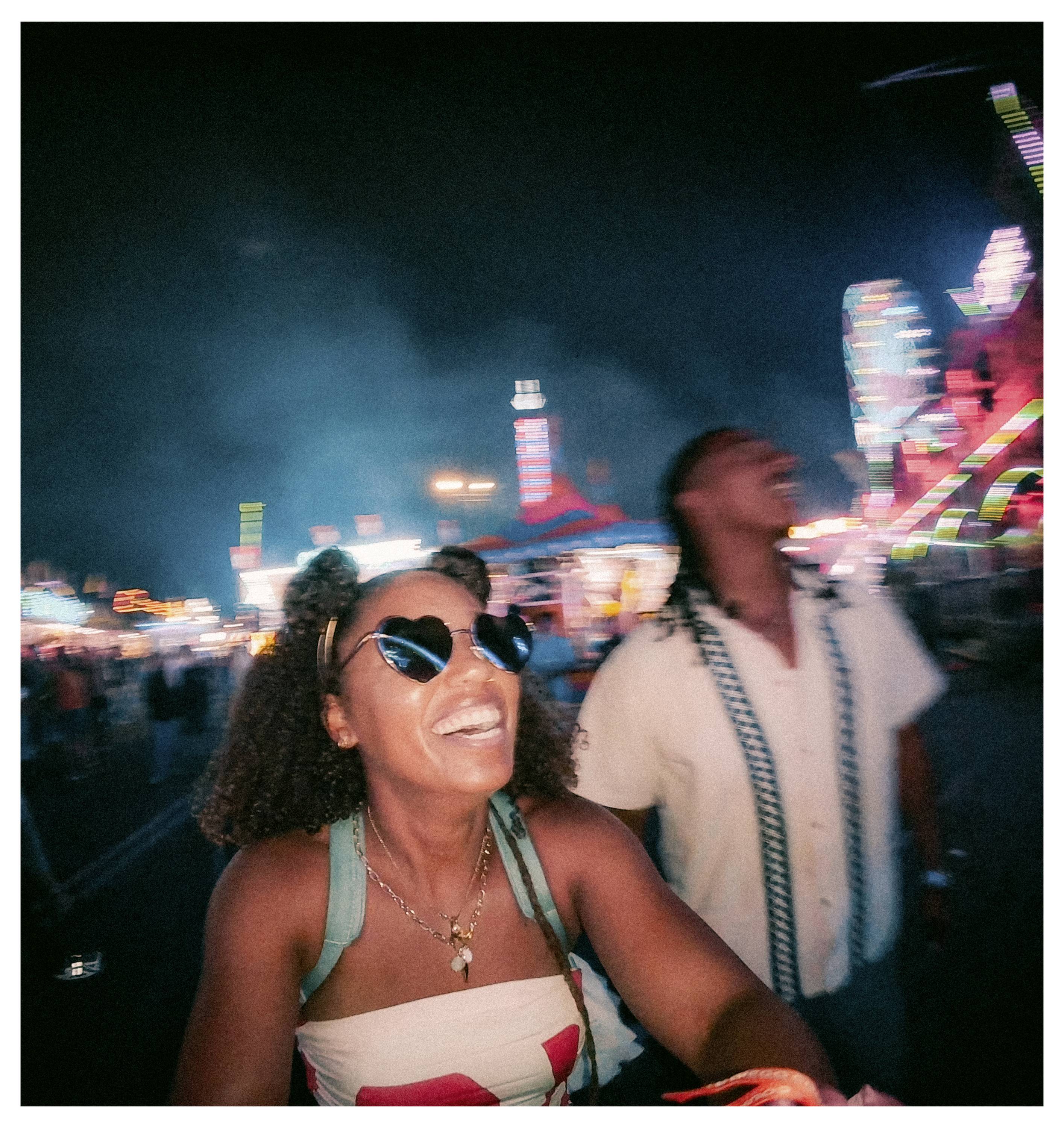 A woman and man enjoy a night at a vibrant carnival, with colorful lights and rides in the background. Both appear joyful and laughing.