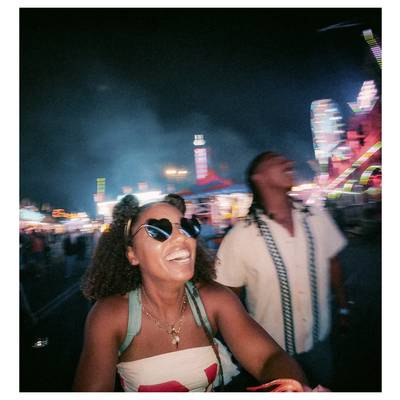 A woman and man enjoy a night at a vibrant carnival, with colorful lights and rides in the background. Both appear joyful and laughing.