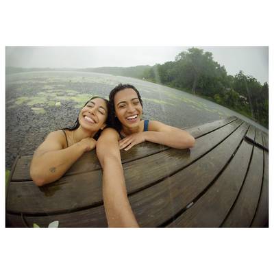 Two smiling people lean on a wooden dock edge, with a lush lake and greenery in the background, capturing a joyful outdoor moment.