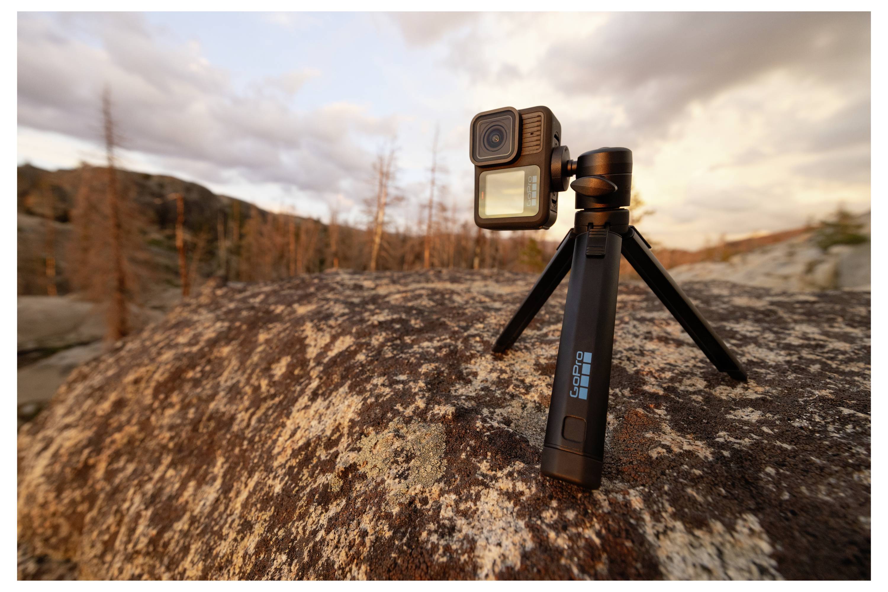 Action camera mounted on a small tripod on a rocky surface in a natural setting, with a scenic view of mountains and trees at sunset.