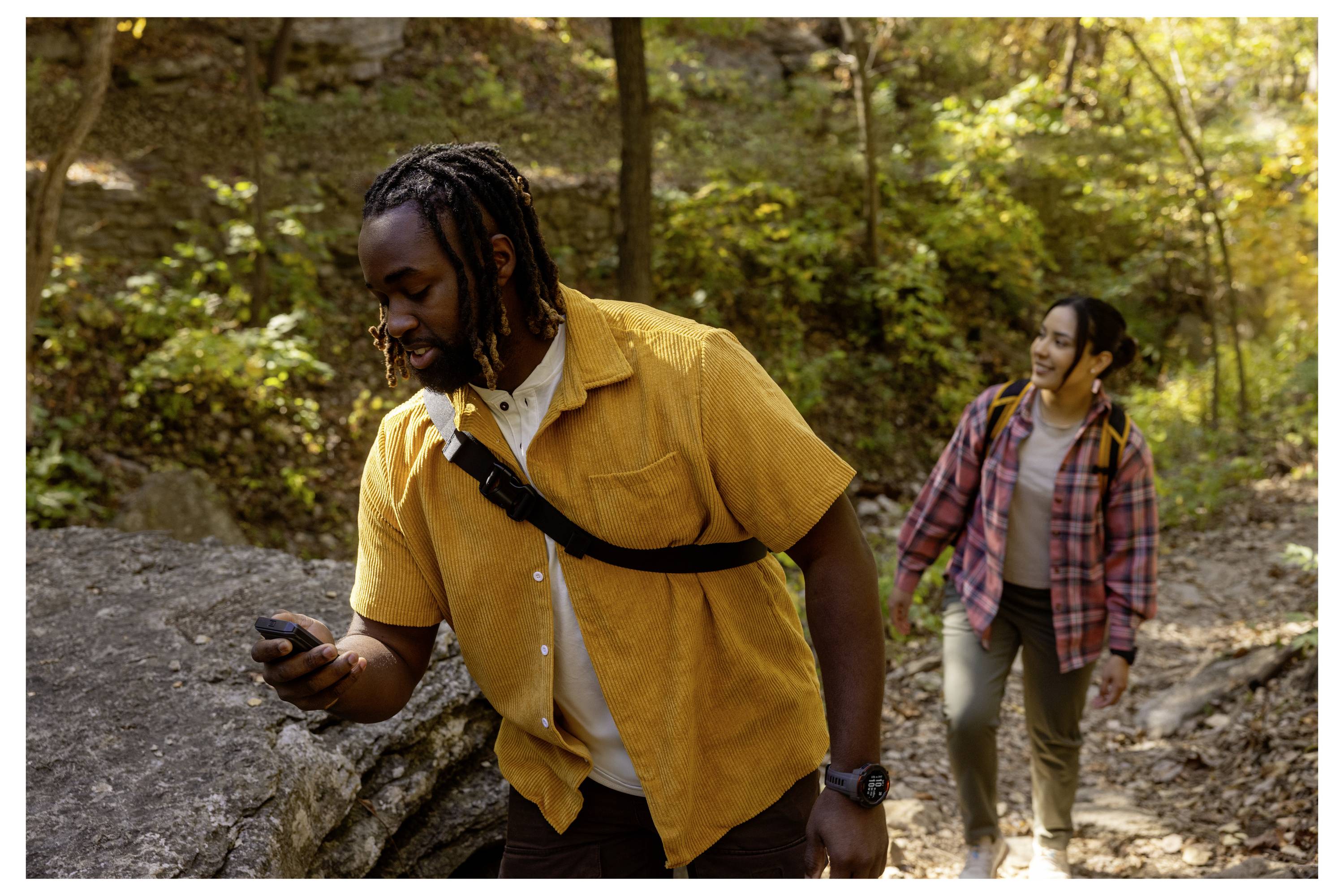 Two people hiking in a forest, one in a yellow shirt holding a GPS device, while the other follows with a smile, wearing a plaid shirt.