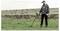 A person using a metal detector on grassy land with a stone wall and cows in the background, suggesting a search or exploration activity.